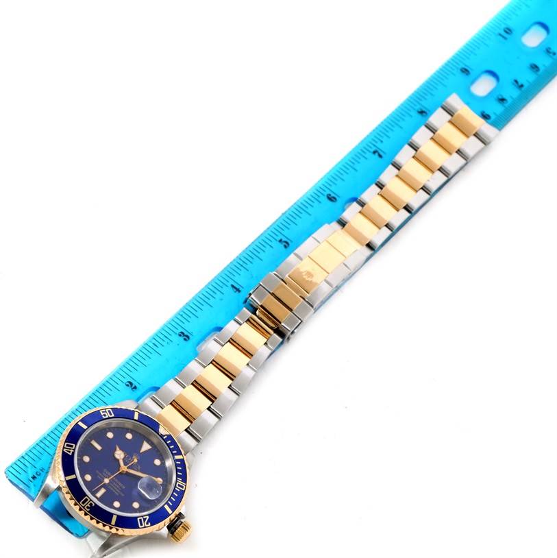 The image shows a Rolex Submariner 16613 Men's Steel and Gold (two tone) Blue Dial 16613 Men's Steel and Gold (two tone) Blue Dial watch with a blue face and gold-and-silver bracelet, laid flat against a blue ruler.