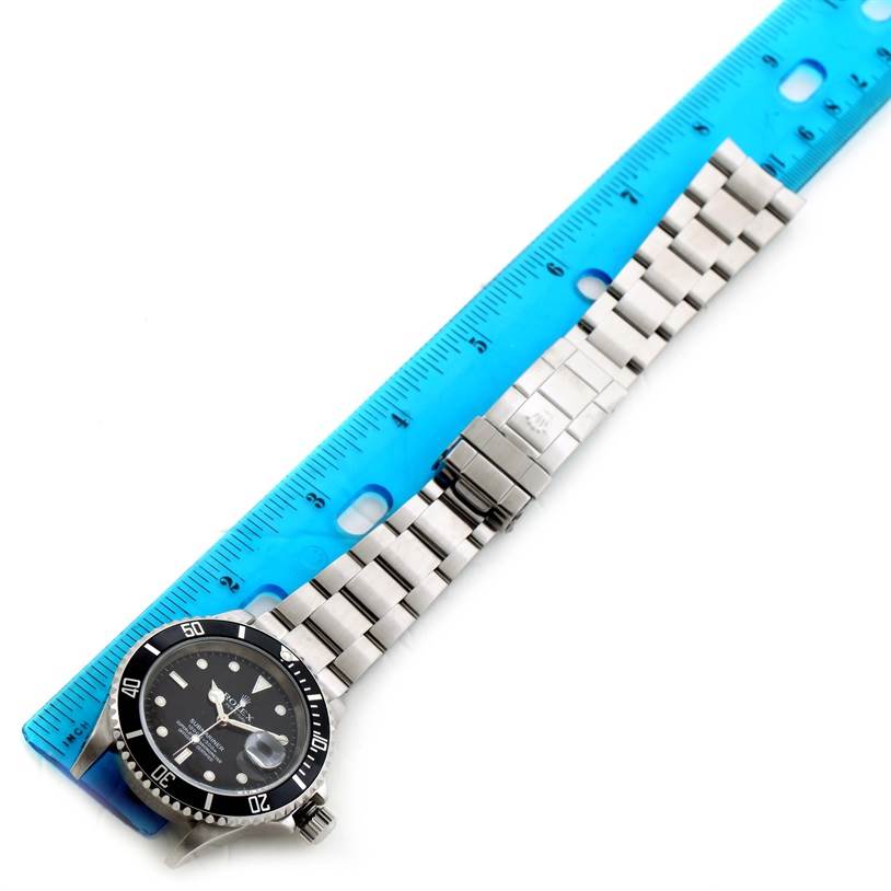 The Rolex Submariner 16610 Men
s Stainless Steel Black Dial 16610 Men
s Stainless Steel Black Dial watch is shown lying flat, alongside a blue ruler to measure its length, displaying the face, bezel, and bracelet.