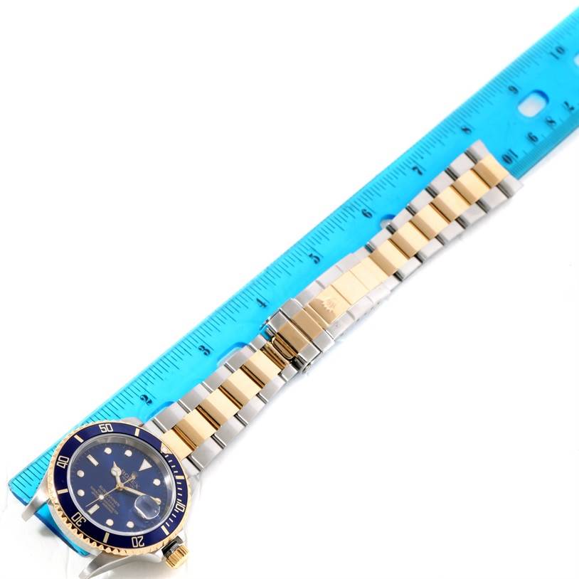 The image shows a Rolex Submariner 16613 Men's Steel and Gold (two tone) Blue Dial 16613 Men's Steel and Gold (two tone) Blue Dial watch laid flat with a blue and gold dial and a gold and silver bracelet next to a blue ruler.