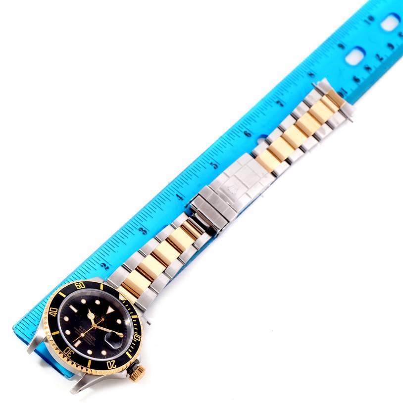 The Rolex Submariner 16613 Men's Steel and Gold (two tone) Black Dial 16613 Men's Steel and Gold (two tone) Black Dial watch is shown from an overhead angle, displaying the face, bezel, and bracelet alongside a blue ruler for scale.