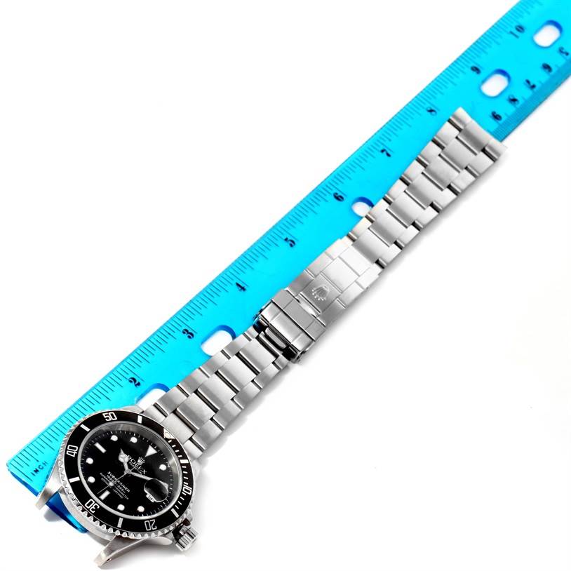 The Rolex Submariner 16610 Men
s Stainless Steel Black Dial 16610 Men
s Stainless Steel Black Dial watch is shown from a top-down angle, highlighting its stainless steel bracelet, clasp, and face next to a ruler.