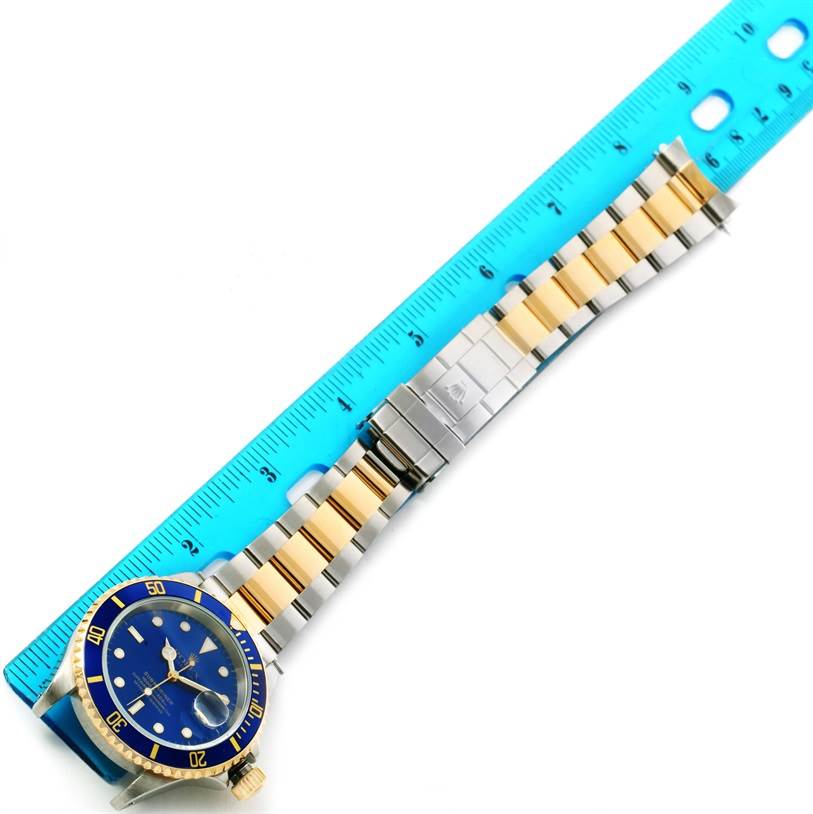 The image shows a Rolex Submariner 16613 Men's Steel and Gold (two tone) Blue Dial 16613 Men's Steel and Gold (two tone) Blue Dial watch laid flat with its stainless steel and gold bracelet next to a blue ruler.