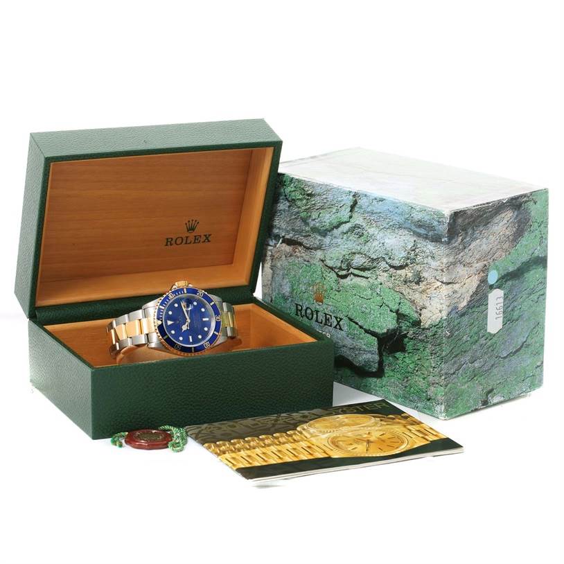 The image shows a Rolex Submariner 16613 Men's Steel and Gold (two tone) Blue Dial 16613 Men's Steel and Gold (two tone) Blue Dial watch in a green and wooden box at a front angle, with its outer box and cards beside it.