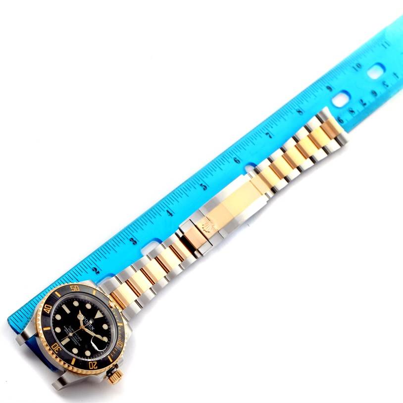 The image shows a Rolex Submariner 116613 Men
s Steel and Gold (two tone) Black Dial 116613 Men
s Steel and Gold (two tone) Black Dial watch laid flat with a focus on its bracelet and bezel next to a blue ruler for scale.