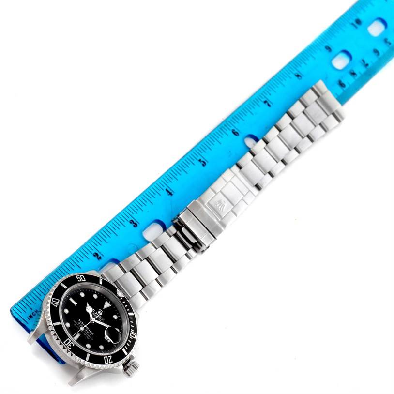 The image shows a Rolex Submariner 16610 Men
s Stainless Steel Black Dial 16610 Men
s Stainless Steel Black Dial watch laid flat alongside a blue ruler, highlighting the watch
s face, bezel, and bracelet.