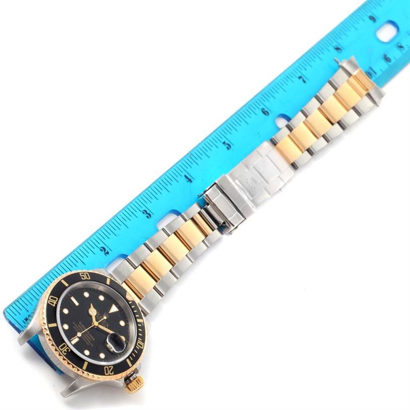 The image shows a Rolex Submariner 16613 Men
s Steel and Gold (two tone) Black Dial 16613 Men
s Steel and Gold (two tone) Black Dial watch laid flat with a blue ruler for scale, highlighting the black dial and gold/silver bracelet.