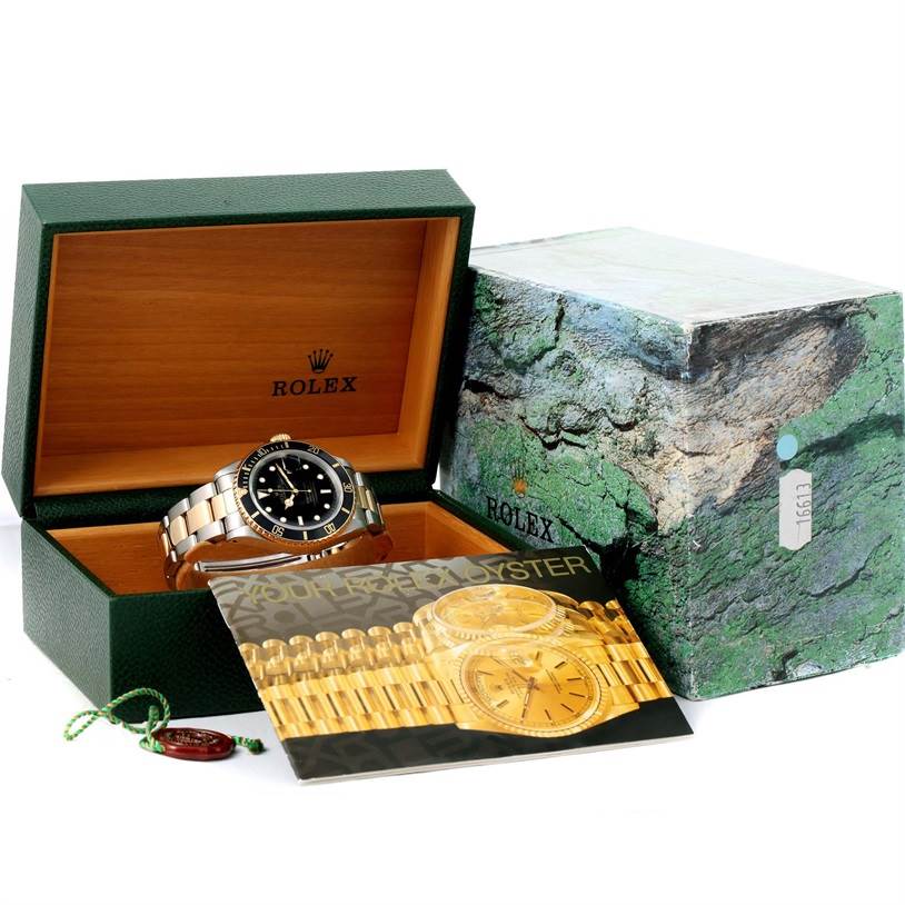 The image shows a Rolex Submariner 16613 Men
s Steel and Gold (two tone) Black Dial 16613 Men
s Steel and Gold (two tone) Black Dial watch in a green box with a booklet, outer packaging, and a tag.