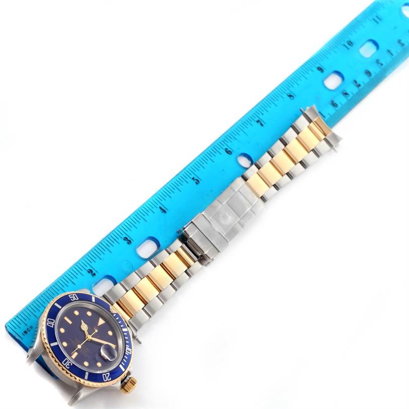 This image shows a Rolex Submariner 16803 Men's Steel and Gold (two tone) Blue Dial 16803 Men's Steel and Gold (two tone) Blue Dial watch lying flat with its blue dial and bracelet band next to a blue ruler for measurement.