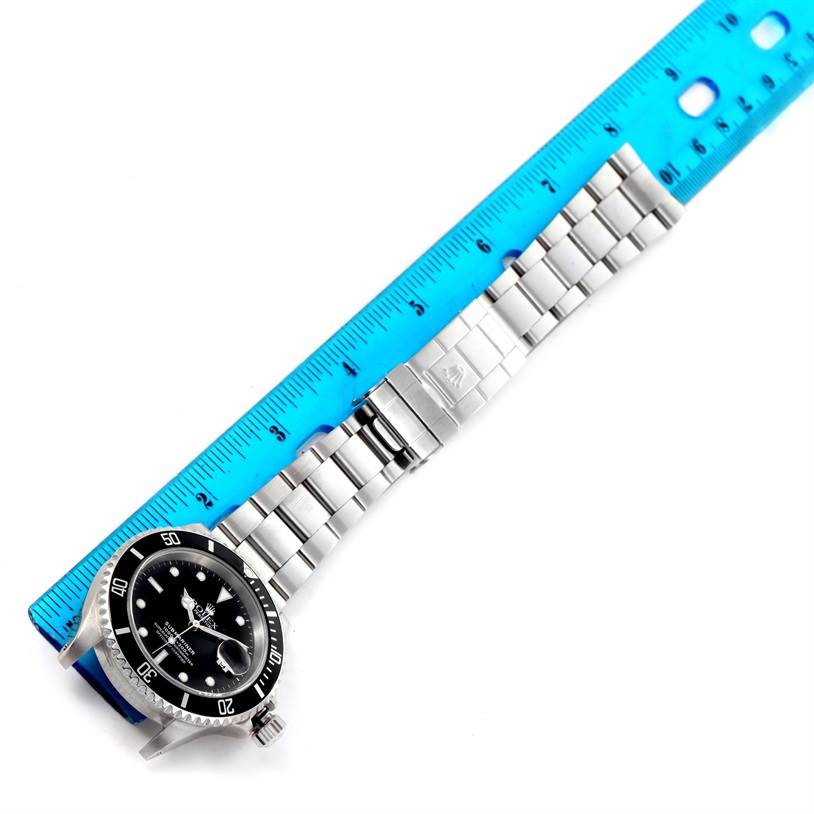 The image shows a Rolex Submariner 16610 Men
s Stainless Steel Black Dial 16610 Men
s Stainless Steel Black Dial watch laid flat with the face and bracelet visible, positioned next to a blue ruler for scale.