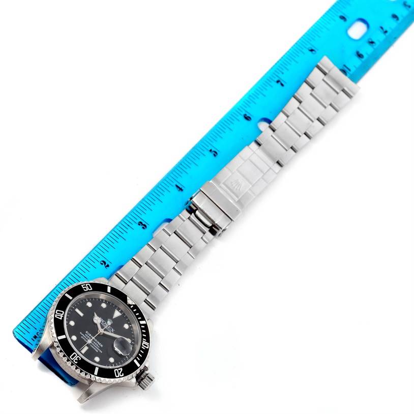 This image shows a Rolex Submariner 16610 Men
s Stainless Steel Black Dial 16610 Men
s Stainless Steel Black Dial watch laid out sideways next to a blue ruler, displaying the watch face, bezel, and metal bracelet.