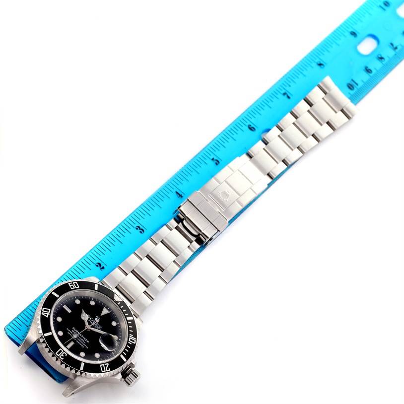 The image shows a Rolex Submariner 16610 Men's Stainless Steel Black Dial 16610 Men's Stainless Steel Black Dial watch lying flat next to a blue ruler, displaying its dial, bezel, and metal bracelet with clasp.