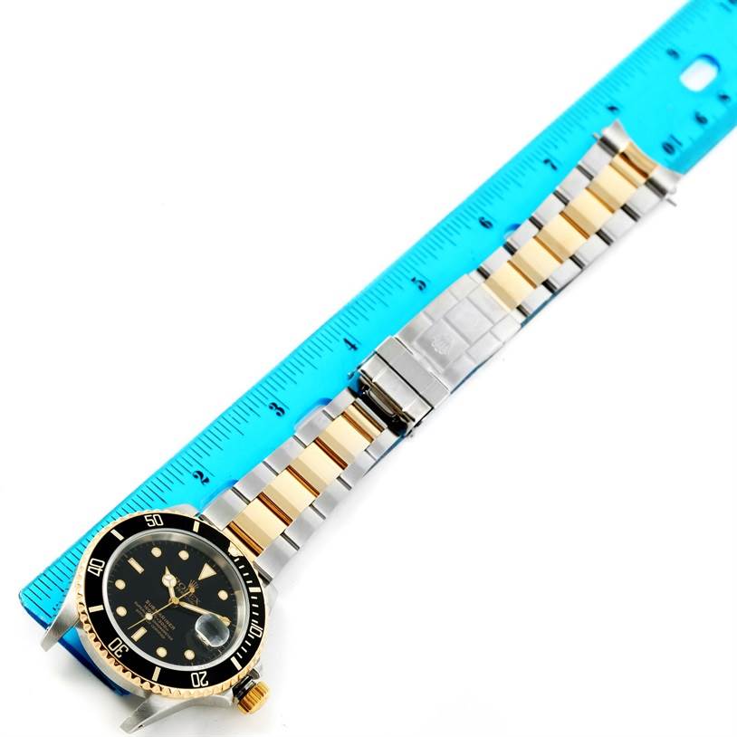 The image shows a top-down view of a Rolex Submariner 16613 Men's Steel and Gold (two tone) Black Dial 16613 Men's Steel and Gold (two tone) Black Dial watch with a gold and silver band, placed next to a ruler.