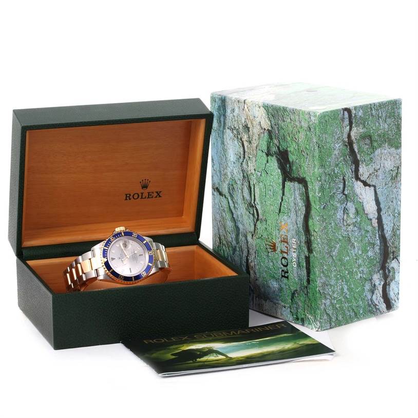 A Rolex Submariner 16613 Men's Steel and Gold (two tone) Grey Dial 16613 Men's Steel and Gold (two tone) Grey Dial watch is displayed in an open green and wood box, with an outer box and booklet placed beside it.