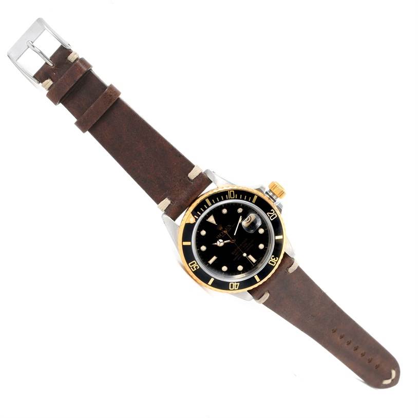 The Rolex Submariner 16803 Men
s Steel and Gold (two tone) Black Dial 16803 Men
s Steel and Gold (two tone) Black Dial watch is shown at an oblique angle, highlighting its black dial, bezel, and brown leather strap.