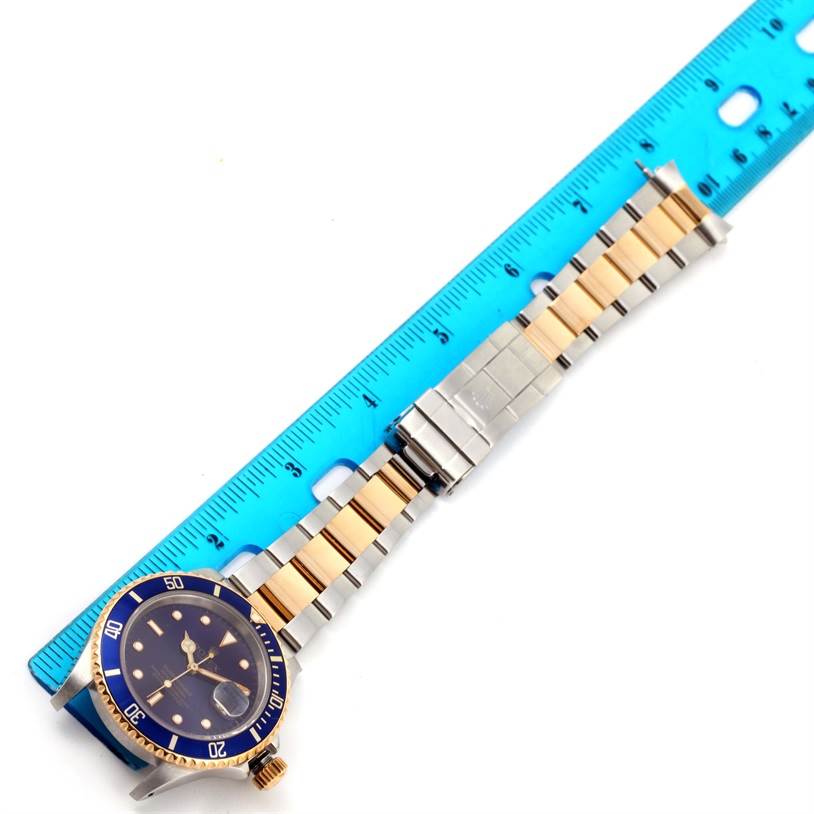 The image shows a Rolex Submariner 16613 Men
s Steel and Gold (two tone) Blue Dial 16613 Men
s Steel and Gold (two tone) Blue Dial watch laid flat, with a blue and gold bezel and a metal bracelet next to a blue ruler for measurement.