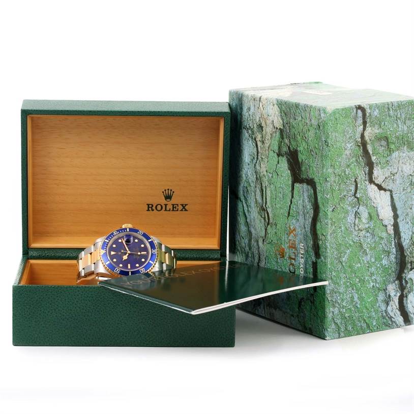 The image shows a Rolex Submariner 16613 Men
s Steel and Gold (two tone) Blue Dial 16613 Men
s Steel and Gold (two tone) Blue Dial watch inside an open box with the outer box and a booklet nearby.
