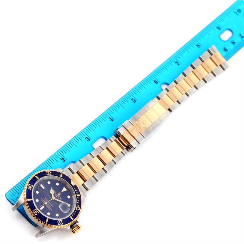 The Rolex Submariner 16613 Men's Steel and Gold (two tone) Blue Dial 16613 Men's Steel and Gold (two tone) Blue Dial watch is shown from a top-down view, including the face and full band, alongside a blue ruler for scale.