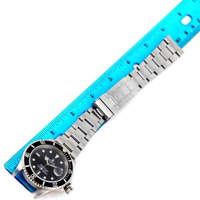 This image displays a Rolex Submariner 16610 Men
s Stainless Steel Black Dial 16610 Men
s Stainless Steel Black Dial watch laid next to a blue ruler, showing the face and metal bracelet.