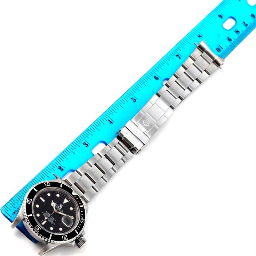 The image shows a Rolex Submariner 168000 Men
s Stainless Steel Black Dial 168000 Men
s Stainless Steel Black Dial watch laid flat with a blue ruler beside it, highlighting the bracelet and dial from an overhead angle.