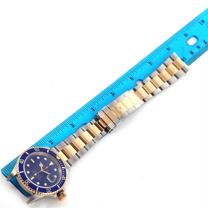 The image shows a Rolex Submariner 16613 Men's Steel and Gold (two tone) Blue Dial 16613 Men's Steel and Gold (two tone) Blue Dial watch laid flat next to a blue ruler, highlighting the bracelet and part of the watch face.