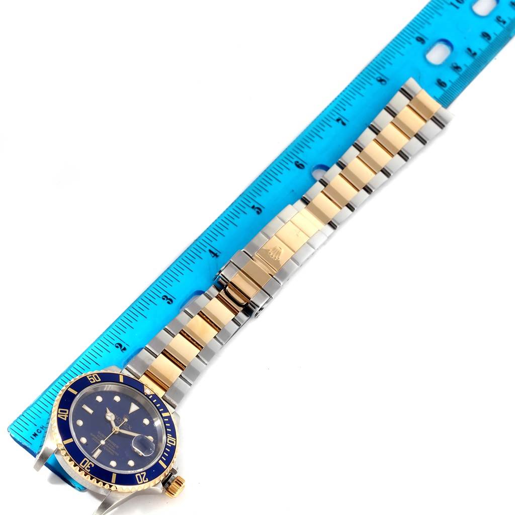 The image shows the Rolex Submariner 16613 Men
s Steel and Gold (two tone) Blue Dial 16613 Men
s Steel and Gold (two tone) Blue Dial watch laid flat beside a blue ruler, displaying its blue dial, bezel, and gold-and-silver bracelet.