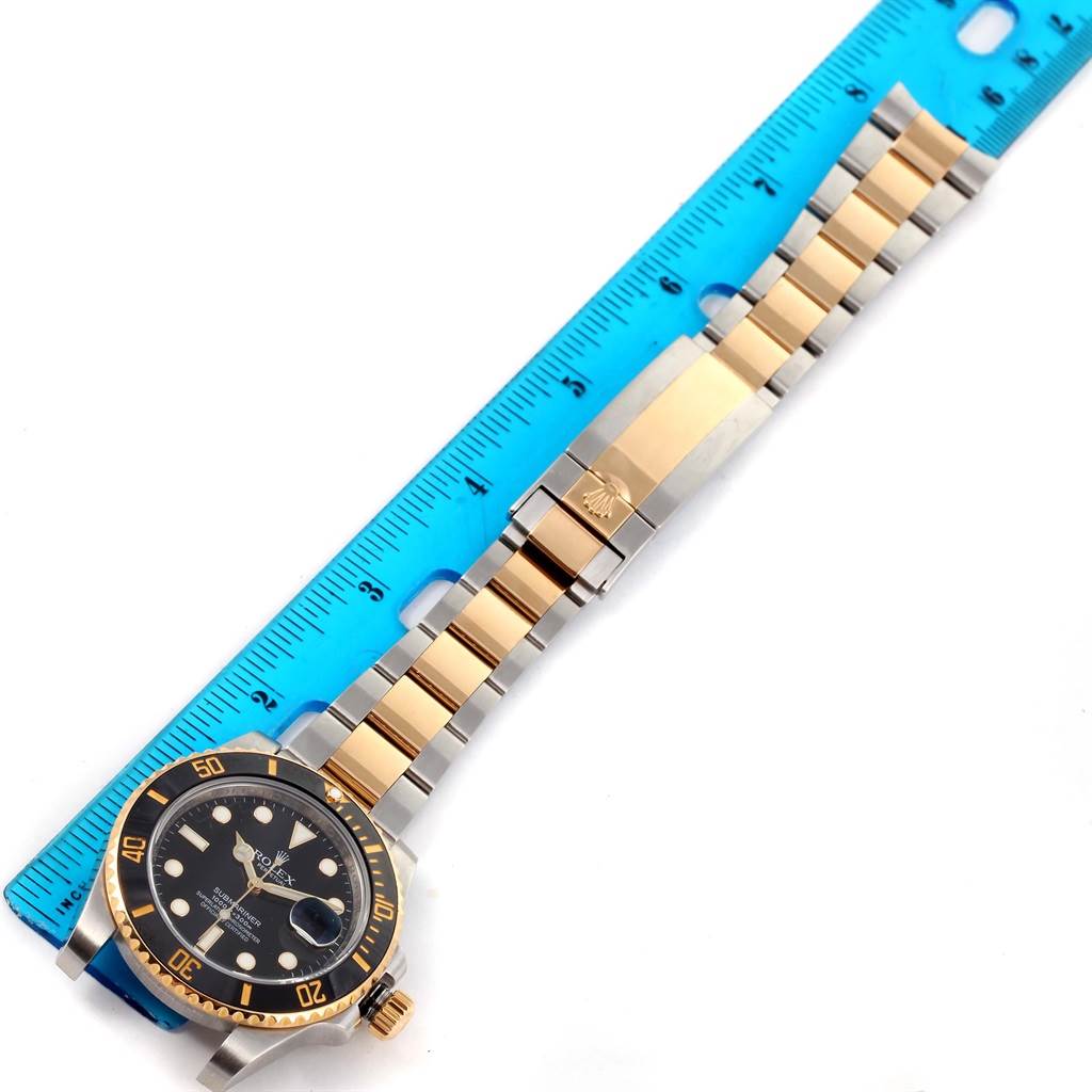 This image shows a Rolex Submariner 116613 Men
s Steel and Gold (two tone) Black Dial 116613 Men
s Steel and Gold (two tone) Black Dial watch with its bracelet extended next to a blue measuring ruler.