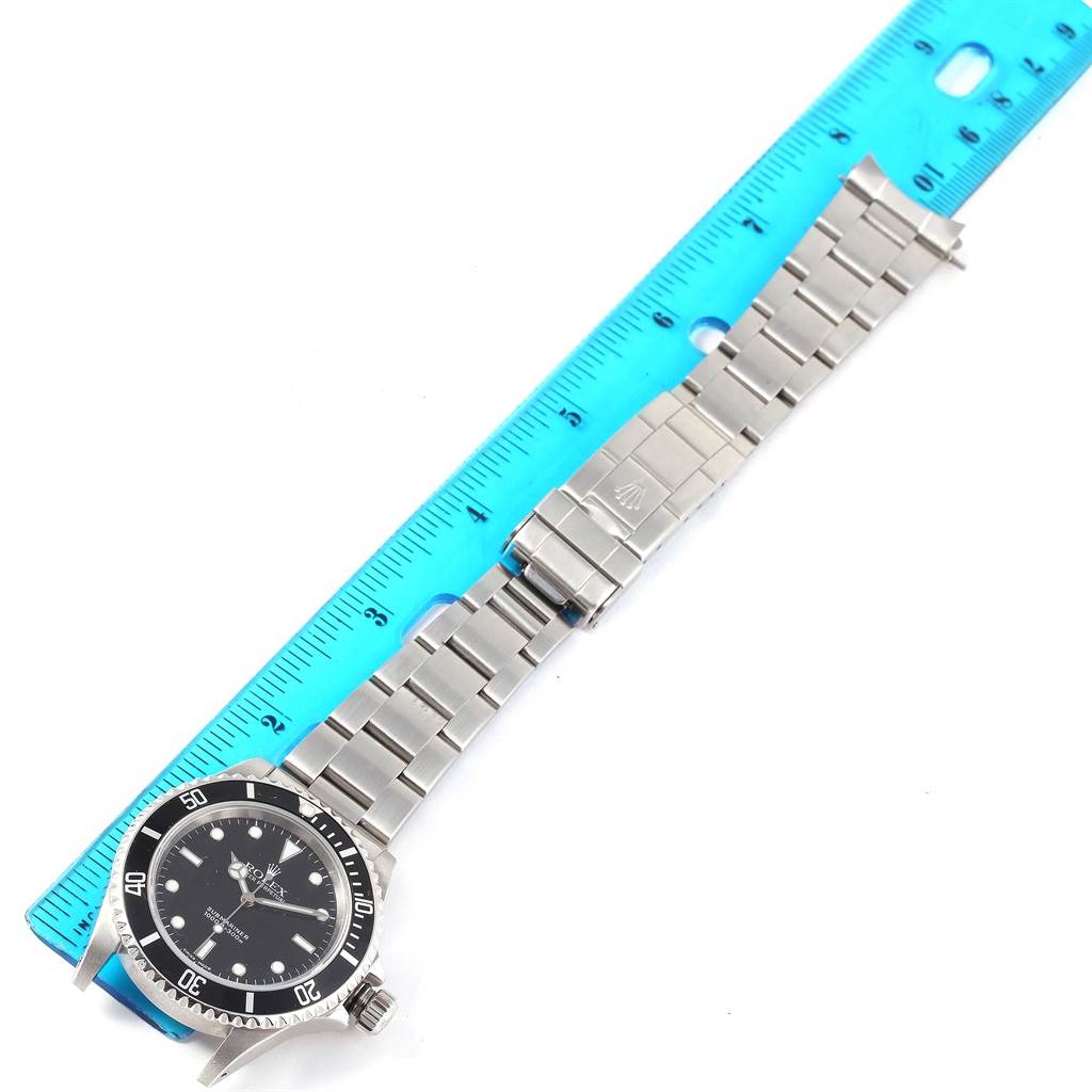 The Rolex Submariner 14060 Men's Stainless Steel Black Dial 14060 Men's Stainless Steel Black Dial watch is shown from a top-down angle with its stainless steel bracelet next to a blue ruler for scale.