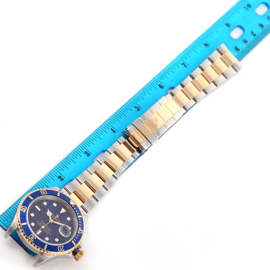 The image shows the Rolex Submariner 16613 Men
s Steel and Gold (two tone) Blue Dial 16613 Men
s Steel and Gold (two tone) Blue Dial watch lying flat alongside a blue ruler, displaying its blue dial, bezel, and two-tone bracelet.