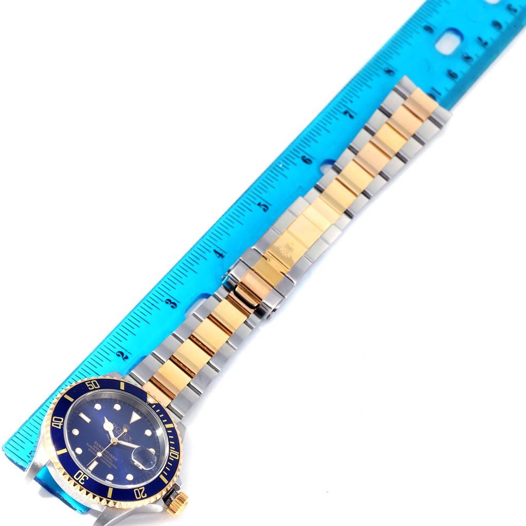 The watch is a Rolex Submariner 16613 Men's Steel and Gold (two tone) Blue Dial 16613 Men's Steel and Gold (two tone) Blue Dial, showing its face, bezel, and metal bracelet beside a blue ruler for scale.