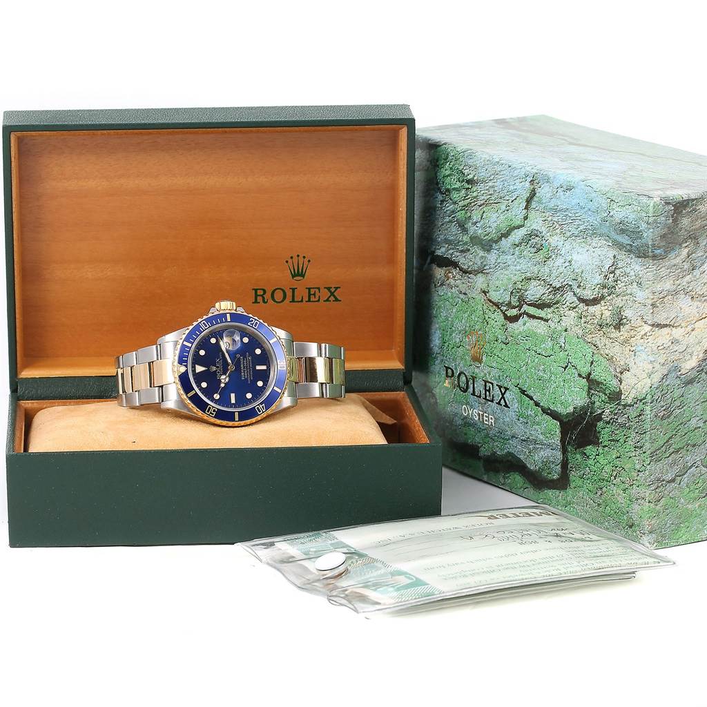 The image shows a Rolex Submariner 16613 Men's Steel and Gold (two tone) Blue Dial 16613 Men's Steel and Gold (two tone) Blue Dial watch with a blue dial inside its open box, along with the outer box and documentation.