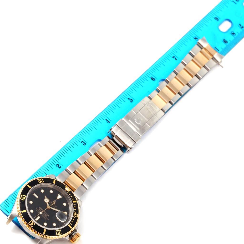 The image shows a top-down view of a Rolex Submariner 16613 Men's Steel and Gold (two tone) Black Dial 16613 Men's Steel and Gold (two tone) Black Dial watch with its band extended alongside a blue ruler.