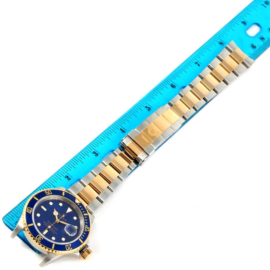 The image shows a Rolex Submariner 16613 Men's Steel and Gold (two tone) Blue Dial 16613 Men's Steel and Gold (two tone) Blue Dial watch laid flat next to a blue ruler, displaying its face, bezel, and two-tone bracelet.