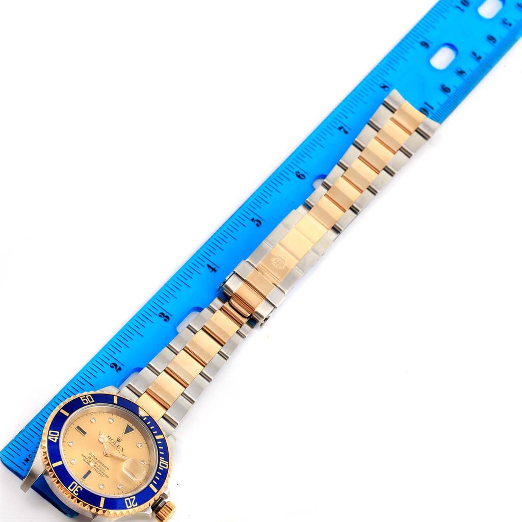 This image shows a Rolex Submariner 16613 Men's Steel and Gold (two tone) Champagne Dial 16613 Men's Steel and Gold (two tone) Champagne Dial watch with a blue and gold bezel, gold dial, and two-tone bracelet, laid next to a ruler.