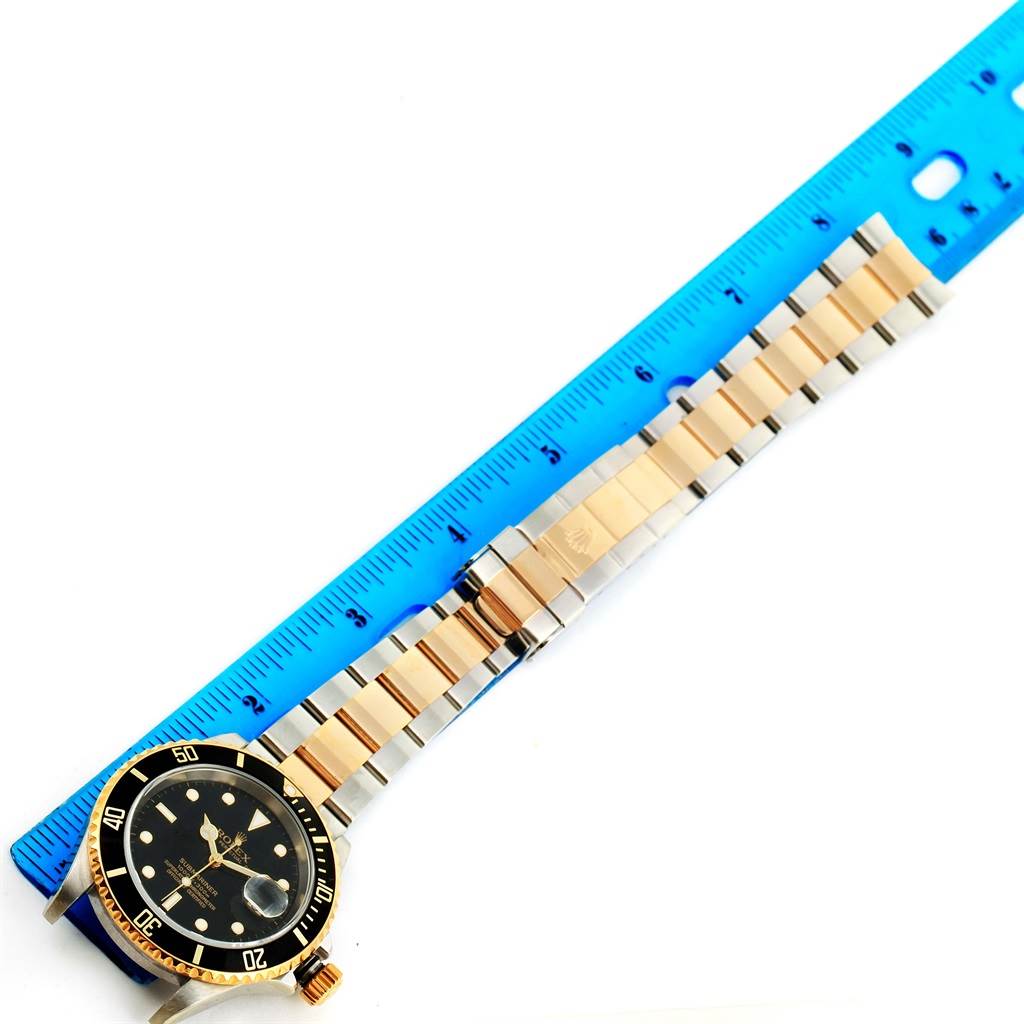 The image shows a Rolex Submariner 16613 Men's Steel and Gold (two tone) Black Dial 16613 Men's Steel and Gold (two tone) Black Dial watch laid flat next to a blue ruler, displaying its face and bracelet.