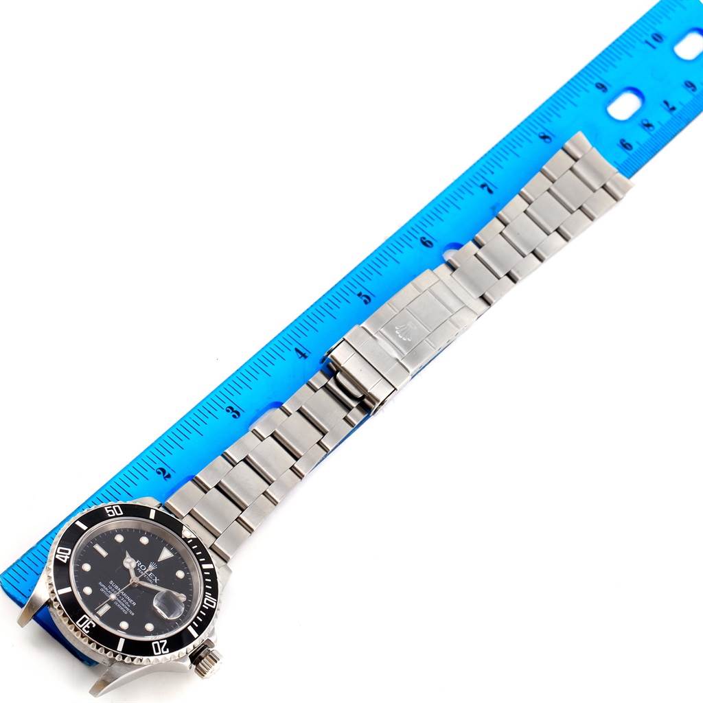 The image shows a Rolex Submariner 16610 Men's Stainless Steel Black Dial 16610 Men's Stainless Steel Black Dial watch laid flat with its stainless steel bracelet fully extended next to a blue ruler.