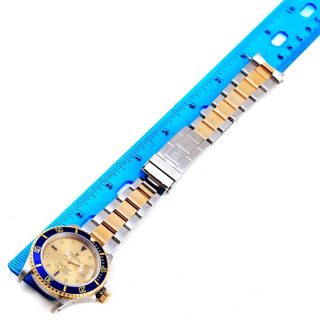 The Rolex Submariner 16613 Men's Steel and Gold (two tone) Champagne Dial 16613 Men's Steel and Gold (two tone) Champagne Dial watch is displayed fully stretched alongside a blue ruler, showing the face and bracelet.