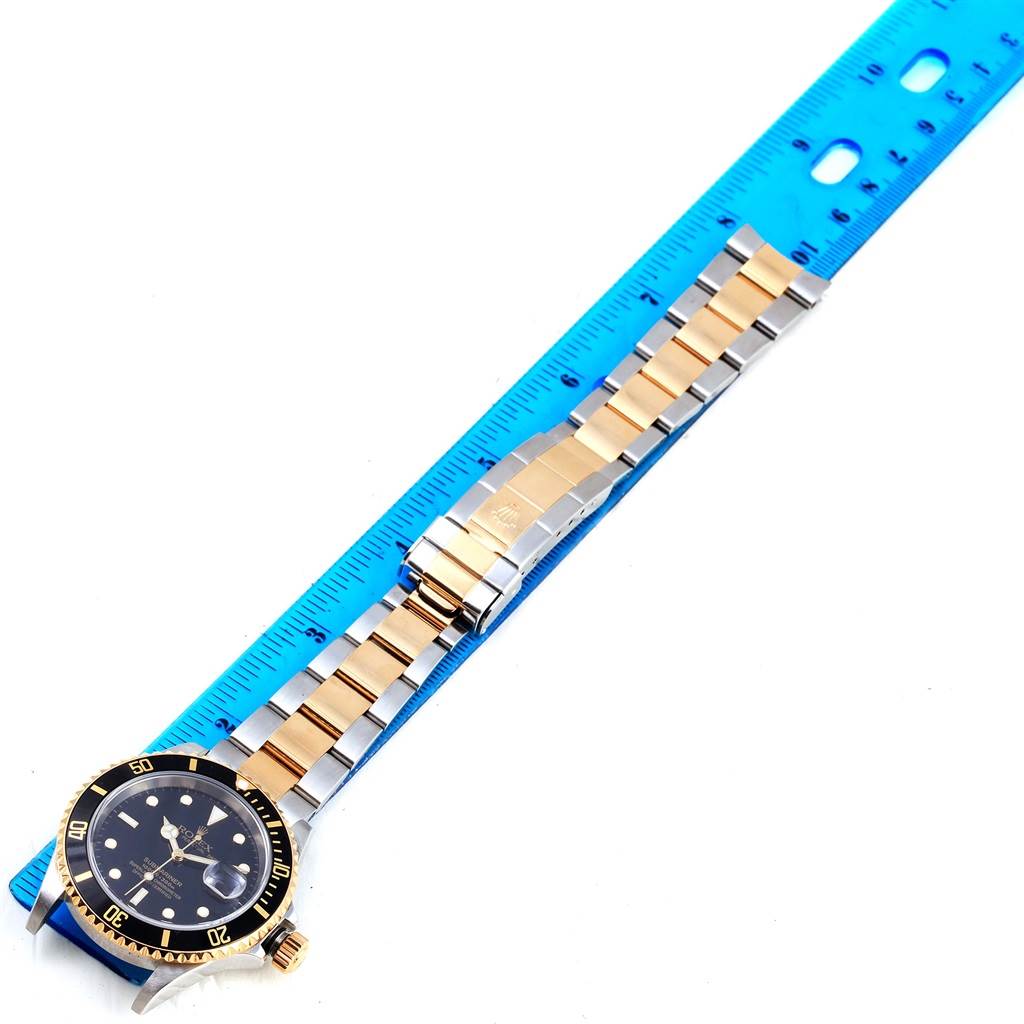 The image shows a Rolex Submariner 16613 Men's Steel and Gold (two tone) Black Dial 16613 Men's Steel and Gold (two tone) Black Dial watch laid flat alongside a blue measuring ruler, highlighting the watch's face and bracelet.