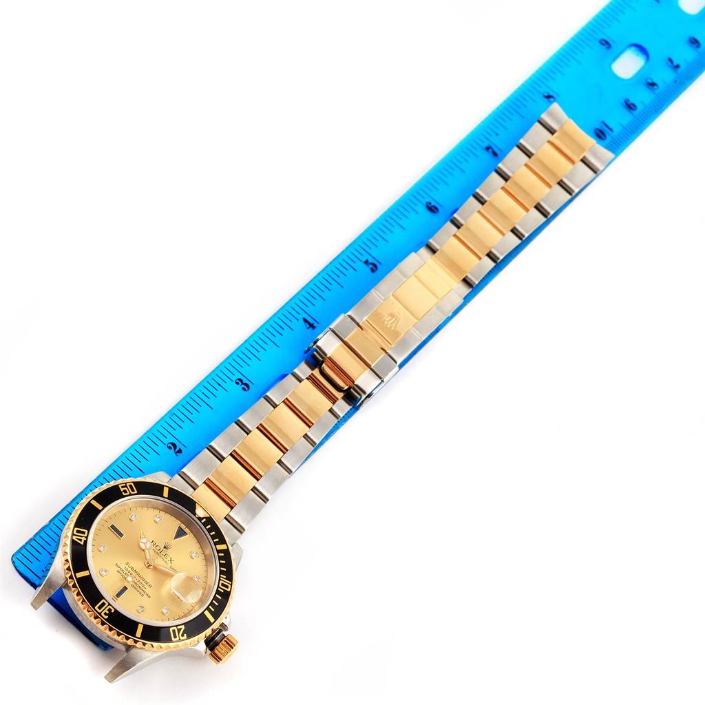The image shows a top view of a Rolex Submariner 16613 Men's Steel and Gold (two tone) Champagne Dial 16613 Men's Steel and Gold (two tone) Champagne Dial watch with a two-tone bracelet beside a blue ruler.
