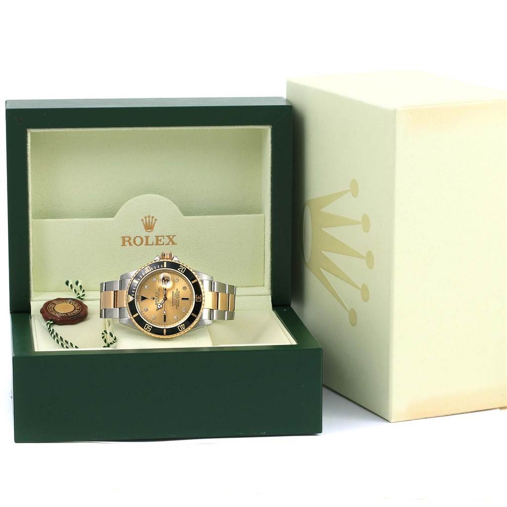 The Rolex Submariner 16613 Men's Steel and Gold (two tone) Champagne Dial 16613 Men's Steel and Gold (two tone) Champagne Dial watch is displayed front-facing in an open green and cream box, with its packaging beside it.