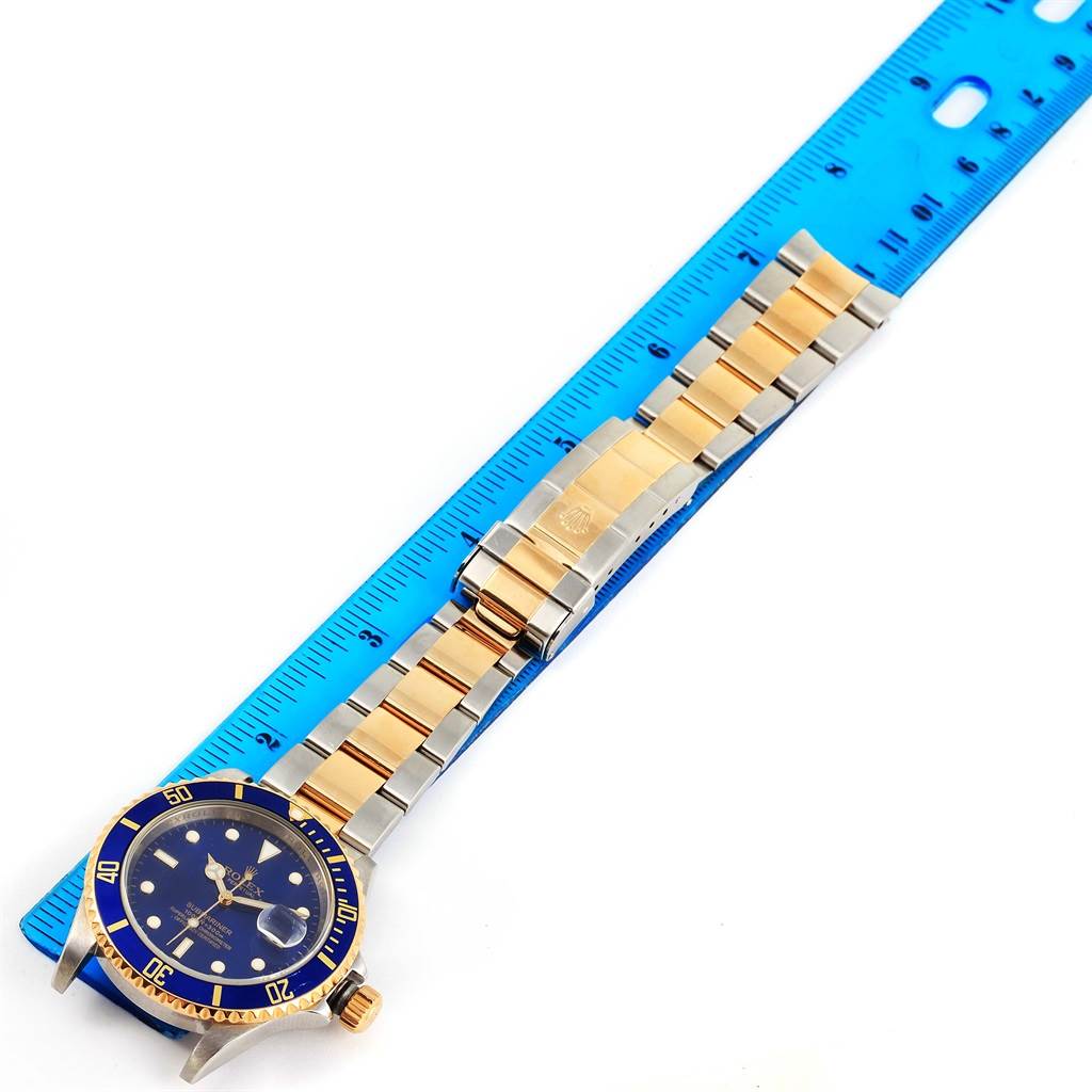 The Rolex Submariner 16613 Men
s Steel and Gold (two tone) Blue Dial 16613 Men
s Steel and Gold (two tone) Blue Dial watch is shown flat with its dial, bezel, and two-tone bracelet visible, placed next to a blue ruler.