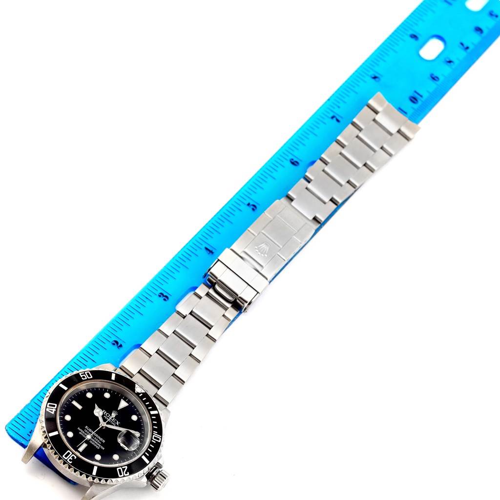 The image shows a Rolex Submariner 16610 Men
s Stainless Steel Black Dial 16610 Men
s Stainless Steel Black Dial watch laid flat next to a ruler, displaying its face, bezel, bracelet, and clasp.