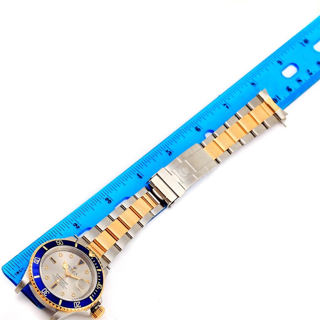 The image shows a Rolex Submariner 16613 Men
s Steel and Gold (two tone) Grey Dial 16613 Men
s Steel and Gold (two tone) Grey Dial watch from a top-down angle, highlighting its bracelet, clasp, and bezel against a ruler for scale.
