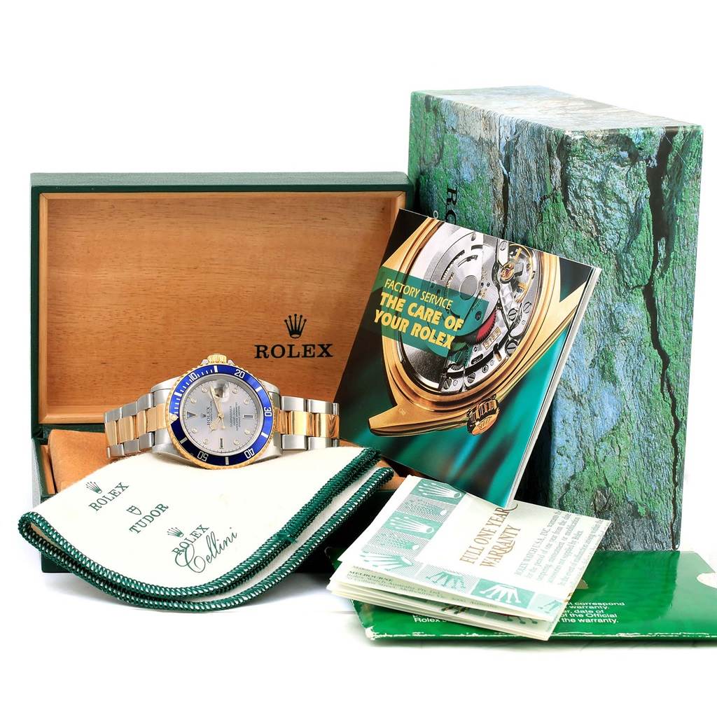 The image shows a Rolex Submariner 16613 Men
s Steel and Gold (two tone) Grey Dial 16613 Men
s Steel and Gold (two tone) Grey Dial with a blue and gold bezel, displayed with its box, warranty card, and care booklet.
