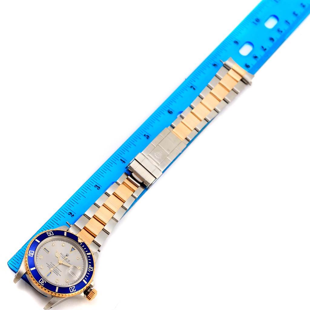 The Rolex Submariner 16613 Men
s Steel and Gold (two tone) Grey Dial 16613 Men
s Steel and Gold (two tone) Grey Dial watch is shown from an overhead angle with its metal bracelet extended alongside a blue ruler.
