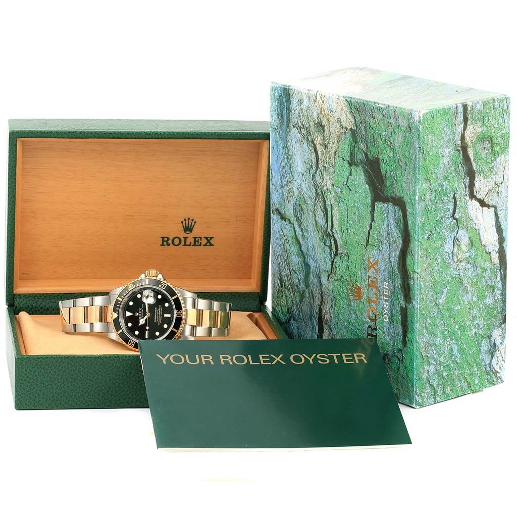 The image shows a Rolex Submariner 16613 Men
s Steel and Gold (two tone) Black Dial 16613 Men
s Steel and Gold (two tone) Black Dial watch, its open wooden box, outer green box, and accompanying booklet from a front angle.