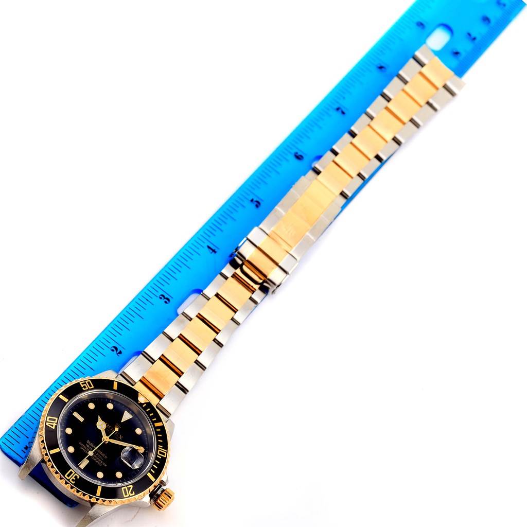 The Rolex Submariner 16613 Men
s Steel and Gold (two tone) Black Dial 16613 Men
s Steel and Gold (two tone) Black Dial watch is shown from a top-down angle, including its face, bezel, and full bracelet, next to a blue ruler.