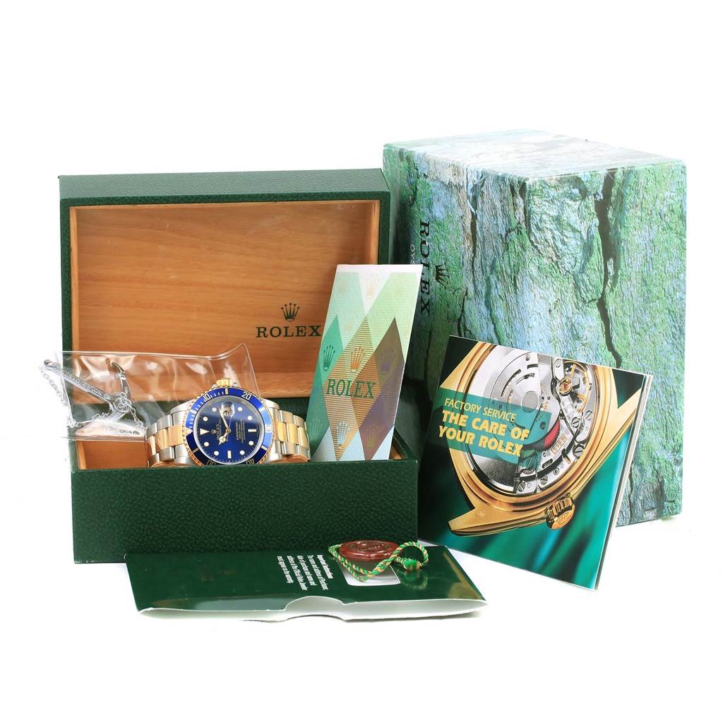 The image shows a Rolex Submariner 16613 Men
s Steel and Gold (two tone) Blue Dial 16613 Men
s Steel and Gold (two tone) Blue Dial watch from the front, resting in its box with accessories, including booklets and tags.