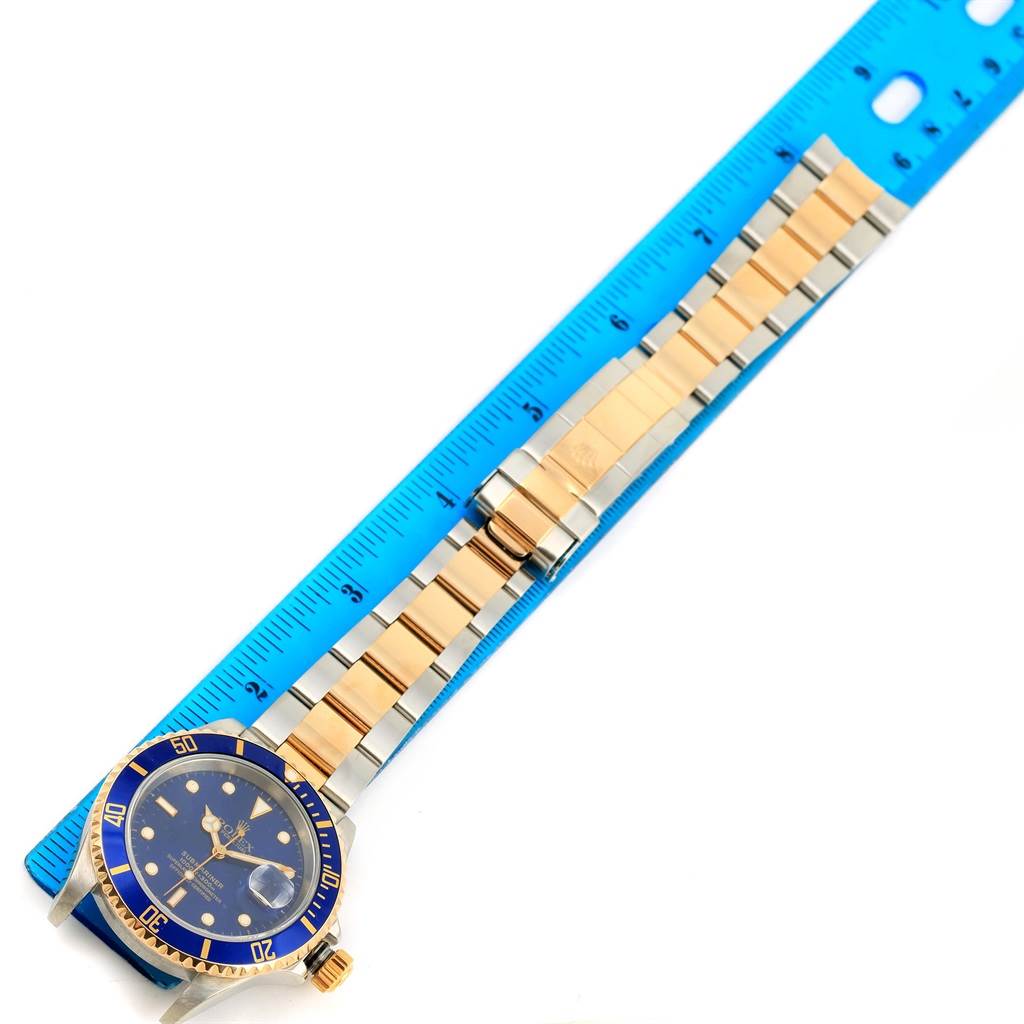 The Rolex Submariner 16613 Men
s Steel and Gold (two tone) Blue Dial 16613 Men
s Steel and Gold (two tone) Blue Dial watch is pictured from a top-down angle, showing the full watch face and the entire band alongside a blue ruler.