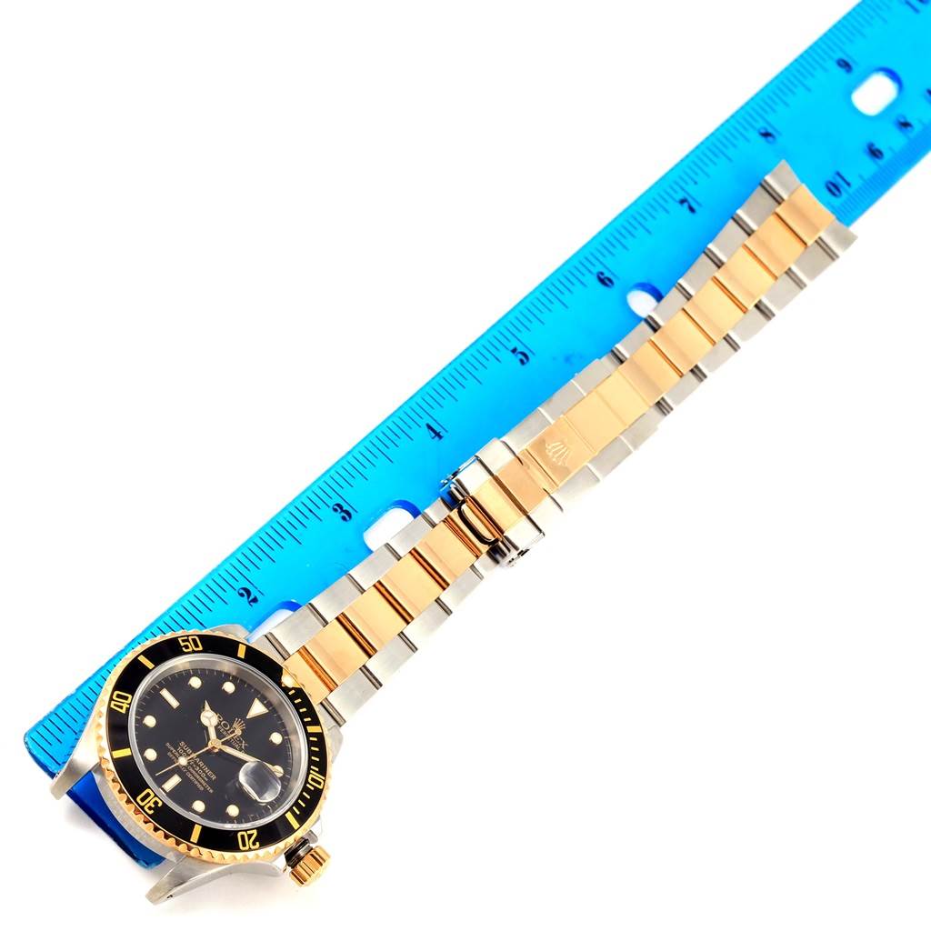 This image shows a Rolex Submariner 16613 Men
s Steel and Gold (two tone) Black Dial 16613 Men
s Steel and Gold (two tone) Black Dial watch lying flat next to a ruler, displaying the watch face, bezel, and strap.