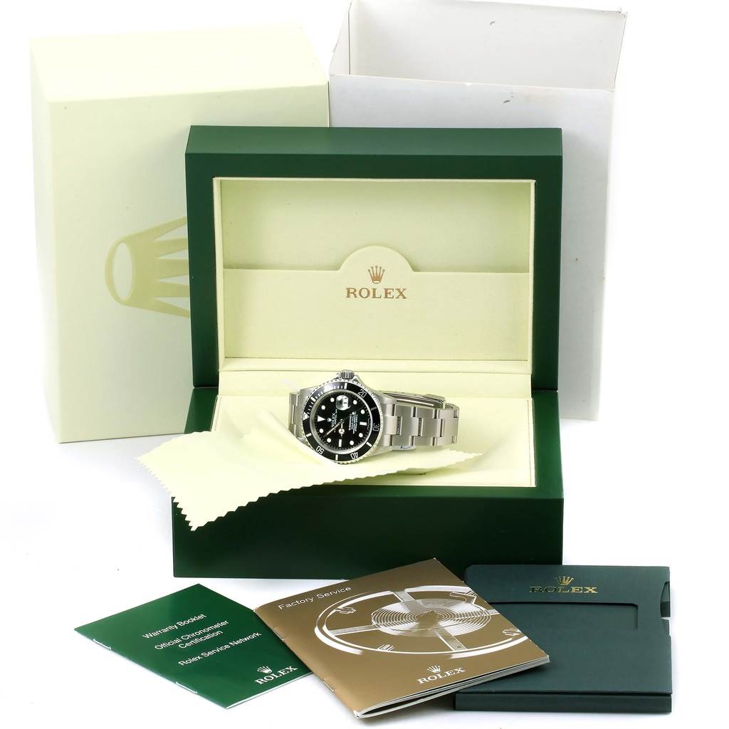 The image shows a Rolex Submariner 16610 Men
s Stainless Steel Black Dial 16610 Men
s Stainless Steel Black Dial watch in its box with accompanying manuals, a cleaning cloth, and an outer box.